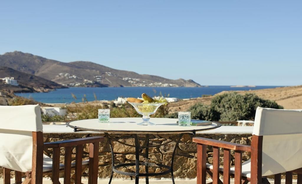 An outdoor dining table with chairs, a bowl of fruit, and glasses, set against a backdrop of a coastal landscape with hills and a view of the sea.