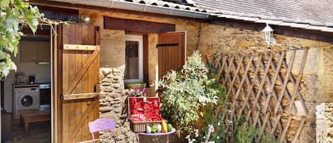 A rustic stone building with a wooden door, a laundry area, and a small garden with a table and chairs.