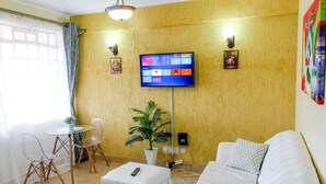 Ein Wohnzimmer mit einer Couch, einem kleinen Tisch, einem Sessel, einer Topfpflanze und einem an der Wand befestigten Fernseher.