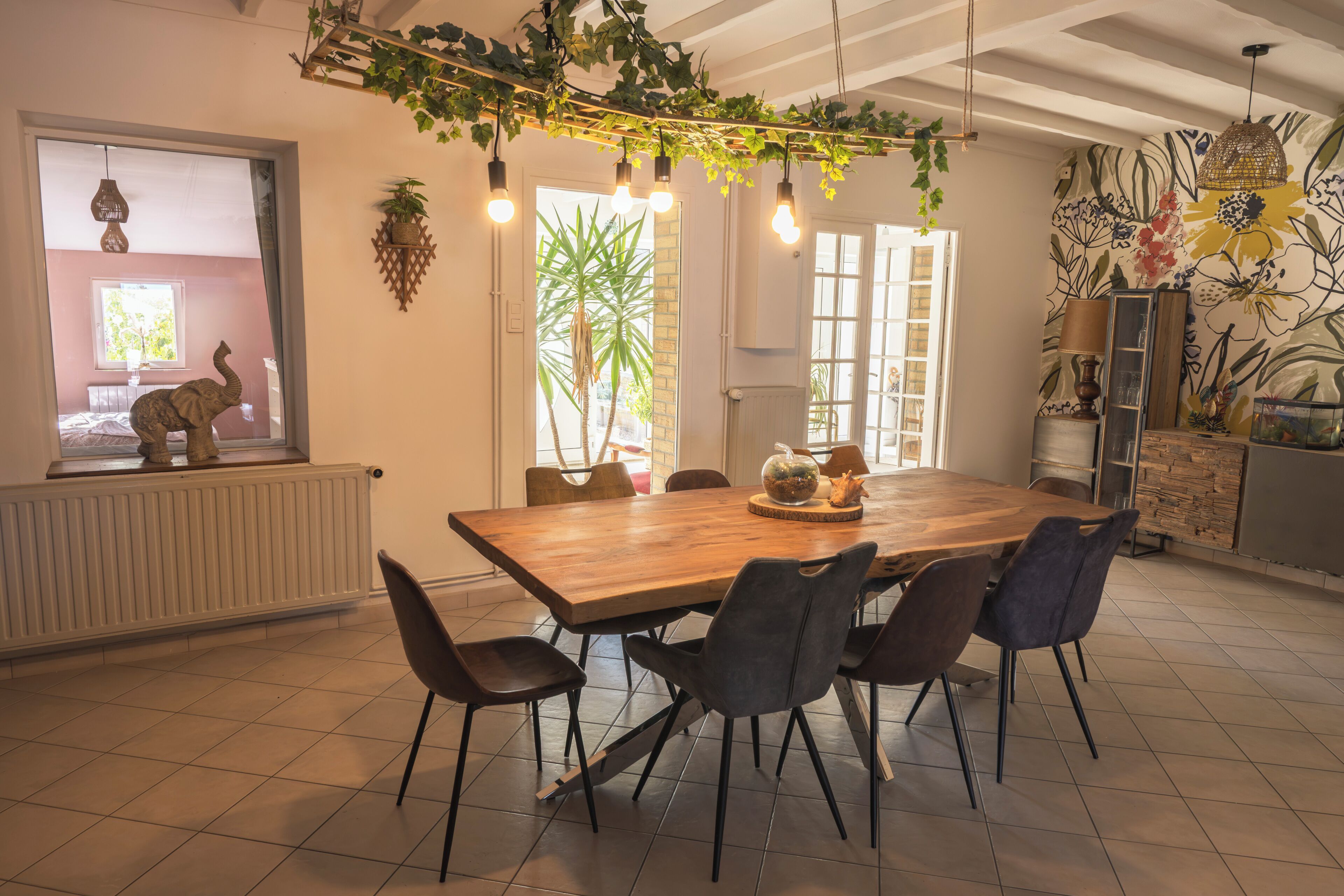 A dining area with a wooden table, six chairs, hanging light bulbs, and a view of a room with a window and a radiator.
