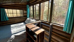 A cabin interior with a bed, a wooden desk, and large windows offering a view of the forest.