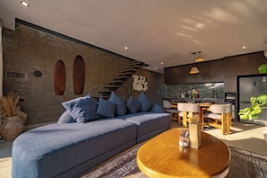 A modern living room with a sofa, coffee table, and kitchen area with a staircase. - Lara Residence 1 Canggu (Canggu)