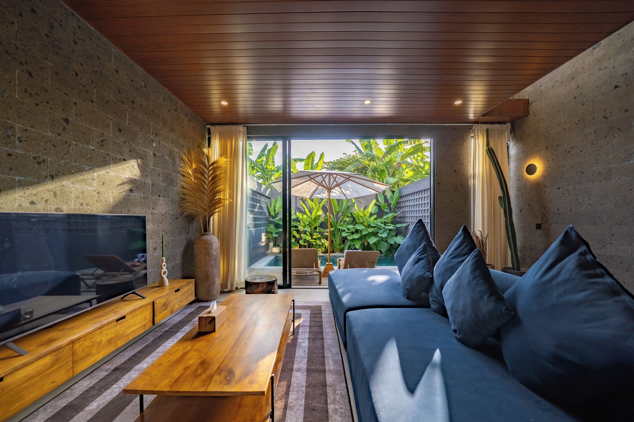 A modern living room with a TV, wooden furniture, a sofa with cushions, and a view of a lush garden through glass doors.