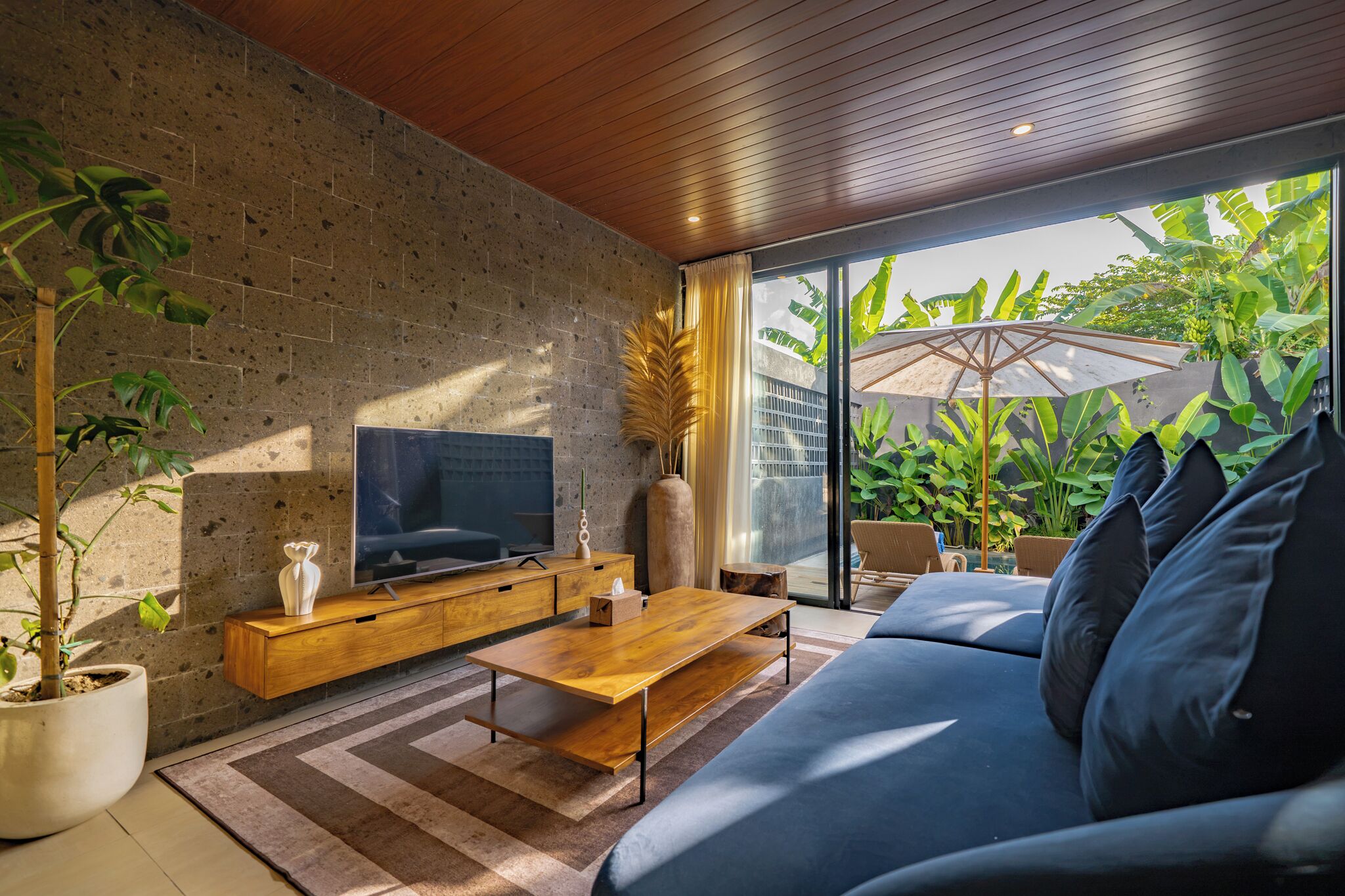 Una sala de estar moderna con pared de piedra, techo de madera, televisor de pantalla plana, sofá y un ventanal con vista a zonas verdes.