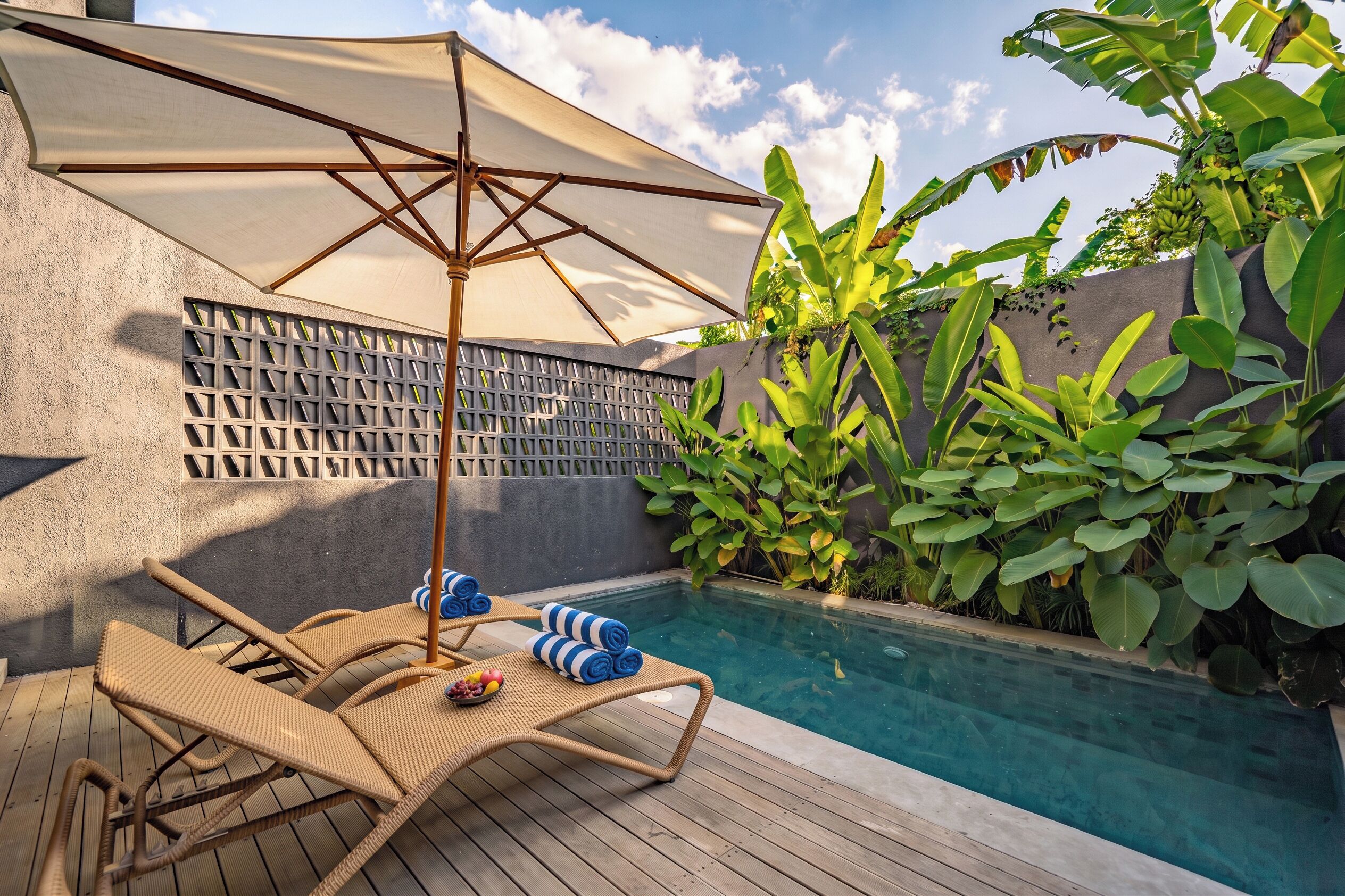 Un área junto a la piscina con sillas de descanso, una sombrilla y plantas tropicales.