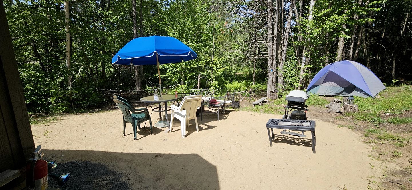 A campsite with a table and chairs, a barbecue grill, a tent, and a blue umbrella.