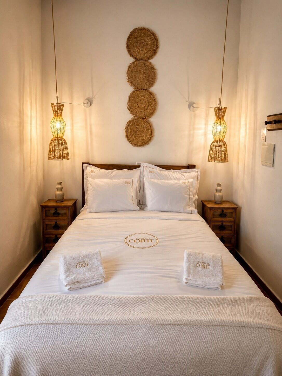 A hotel room with a bed, two bedside tables, a wall-mounted light fixture, and a decorative wall hanging.