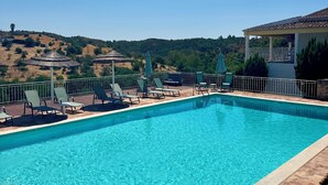 A rectangular swimming pool with clear blue water, surrounded by a wooden deck and metal railings. There are several lounge chairs and umbrellas along the poolside.