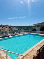 A rectangular swimming pool with clear blue water, surrounded by a wooden deck and metal railings. There are several lounge chairs and umbrellas along the poolside.