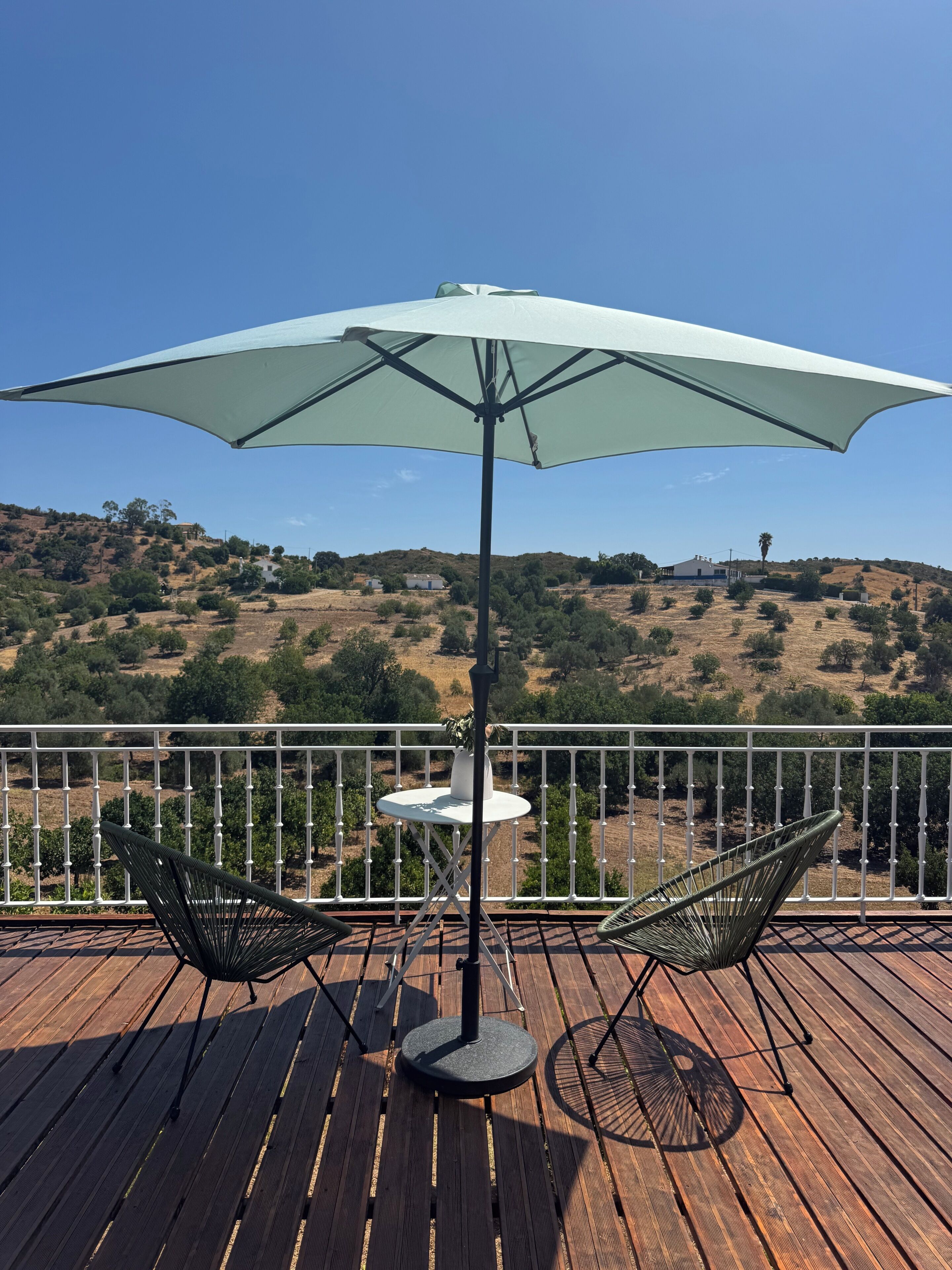 Terraza de madera con dos sillas y una mesa bajo un amplio paraguas, con vistas a un paisaje montañoso.