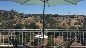 Terraza de madera con dos sillas y una mesa bajo un amplio paraguas, con vistas a un paisaje montañoso.
