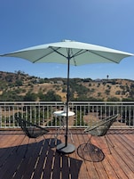 Um deck de madeira com duas cadeiras e uma mesa sob um guarda-sol grande, com vista para uma paisagem ondulada.