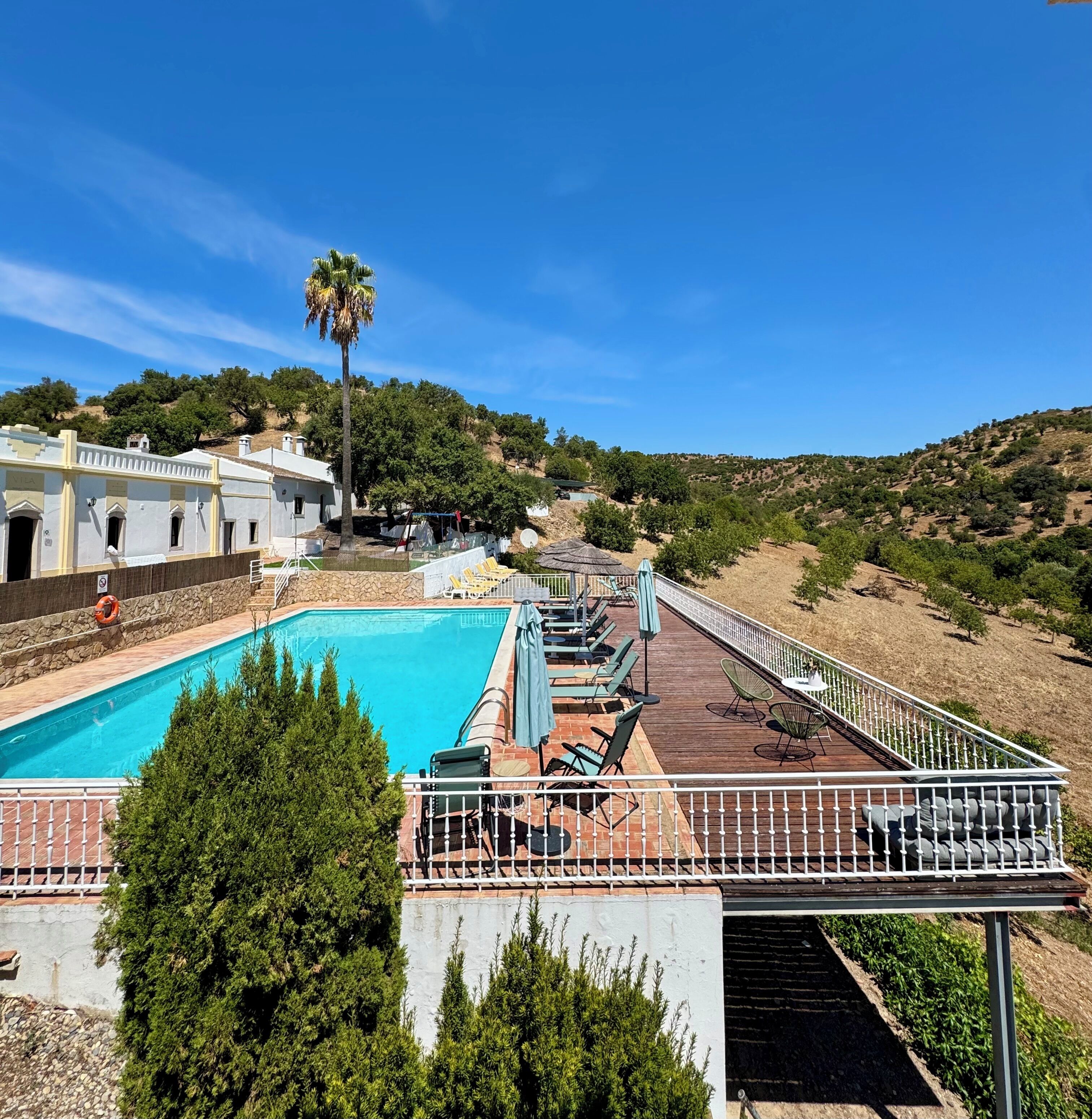 Un área de piscina con terraza de madera, sillas de descanso y vistas a un paisaje montañoso.
