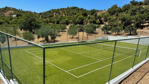 A tennis court with a green surface, surrounded by a chain-link fence, and a hilly landscape in the background.