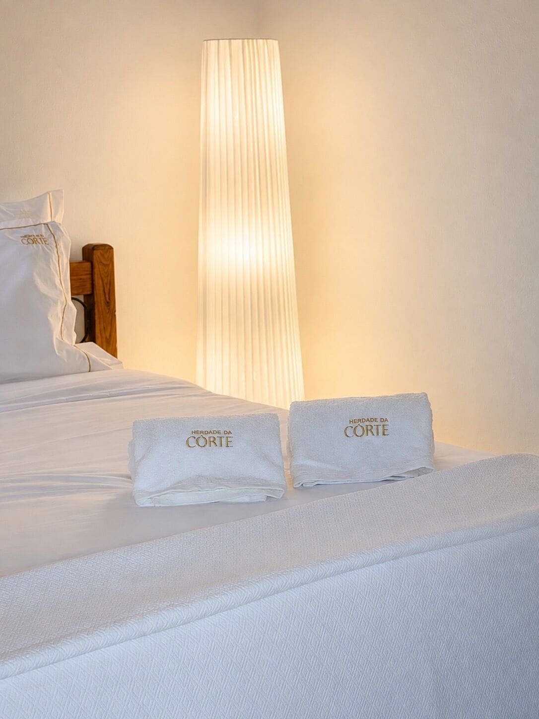 Een keurig opgemaakt bed met twee handdoeken erop, een hoge, verticale lamp en een houten hoofdbord.