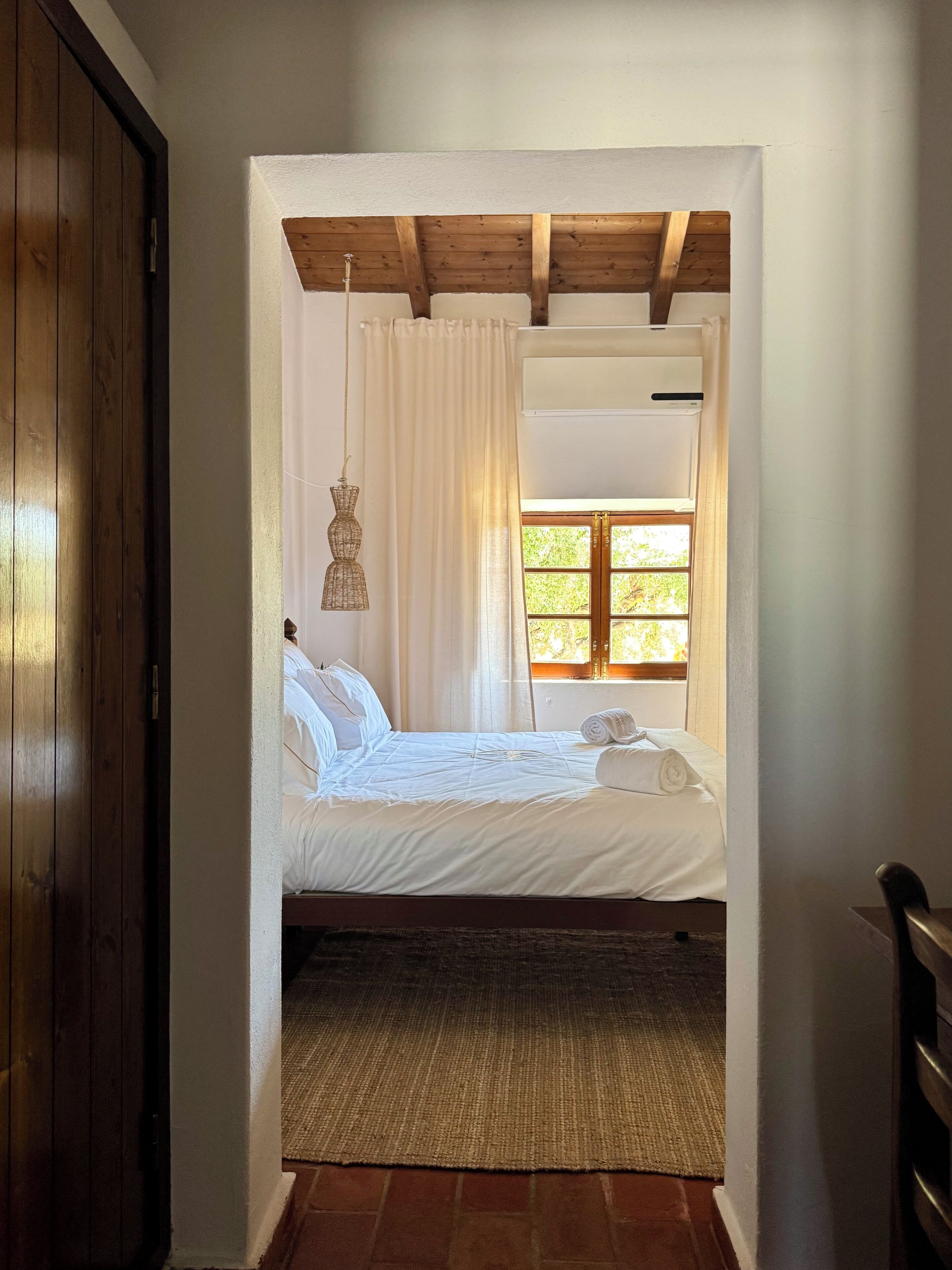 A bedroom with a wooden ceiling, a bed with white bedding, a window with curtains, and a hanging light fixture.