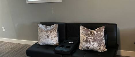 Sala de estar con un sofá de cuero negro, dos almohadones estampados, un control remoto sobre el sofá, una ventana con persianas y un cuadro en la pared.