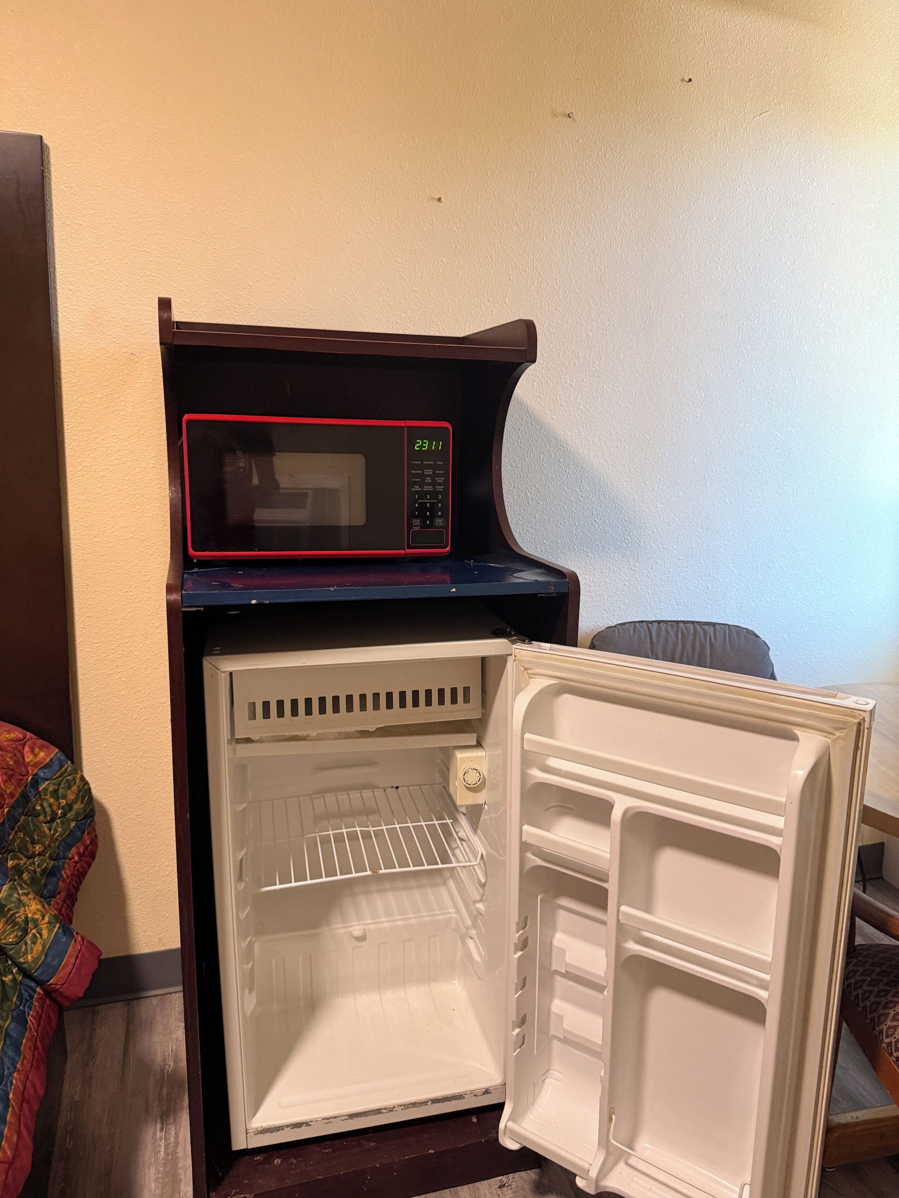A microwave on top of a refrigerator in a room with a plain wall and a patterned cushion.