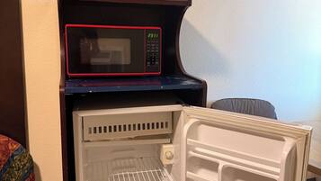 A microwave on top of a refrigerator in a room with a plain wall and a patterned cushion.