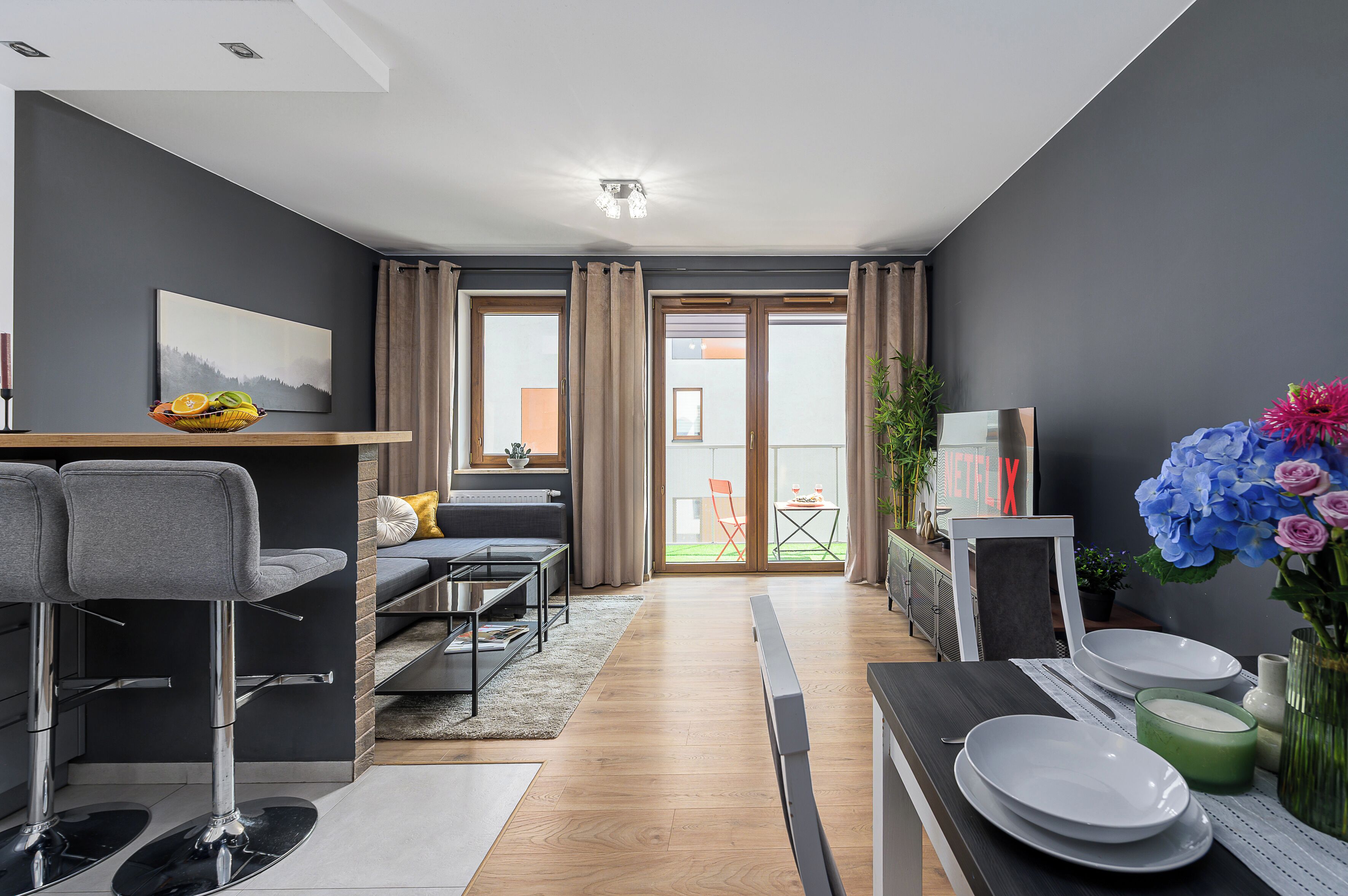 Ein modernes Wohnzimmer mit einer Couch, einem Esstisch mit Tellern, einem Fernseher und Blick ins Freie.