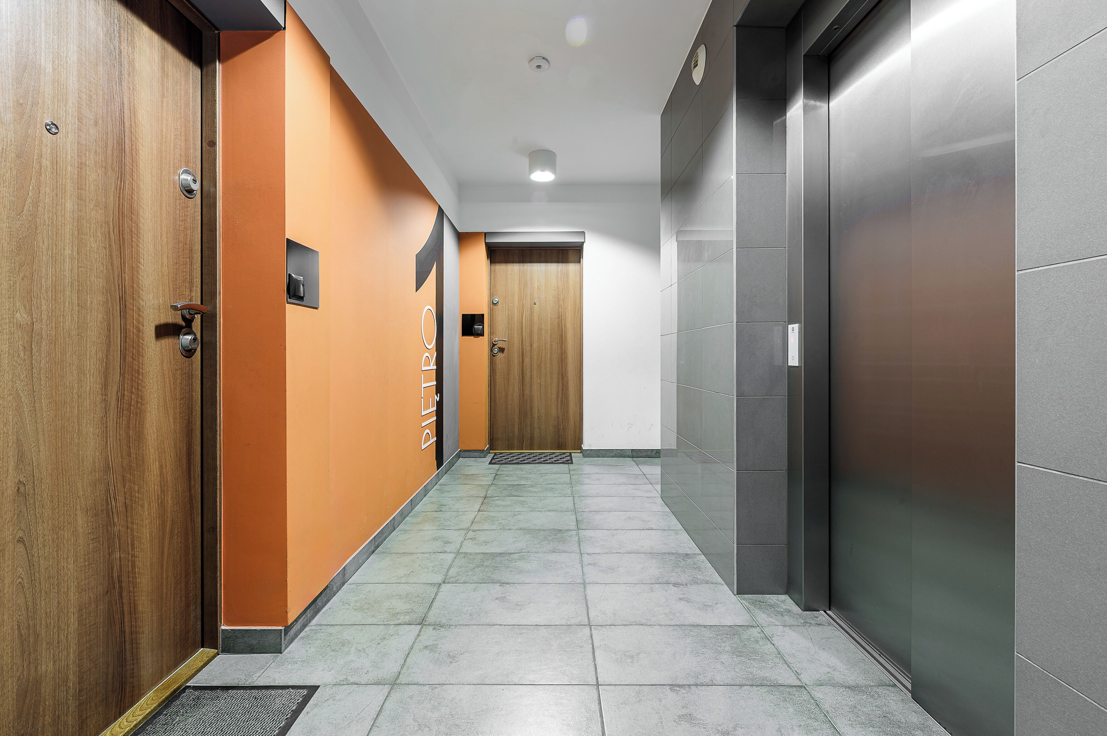 Un couloir avec une porte en bois, un mur orange et un ascenseur gris.