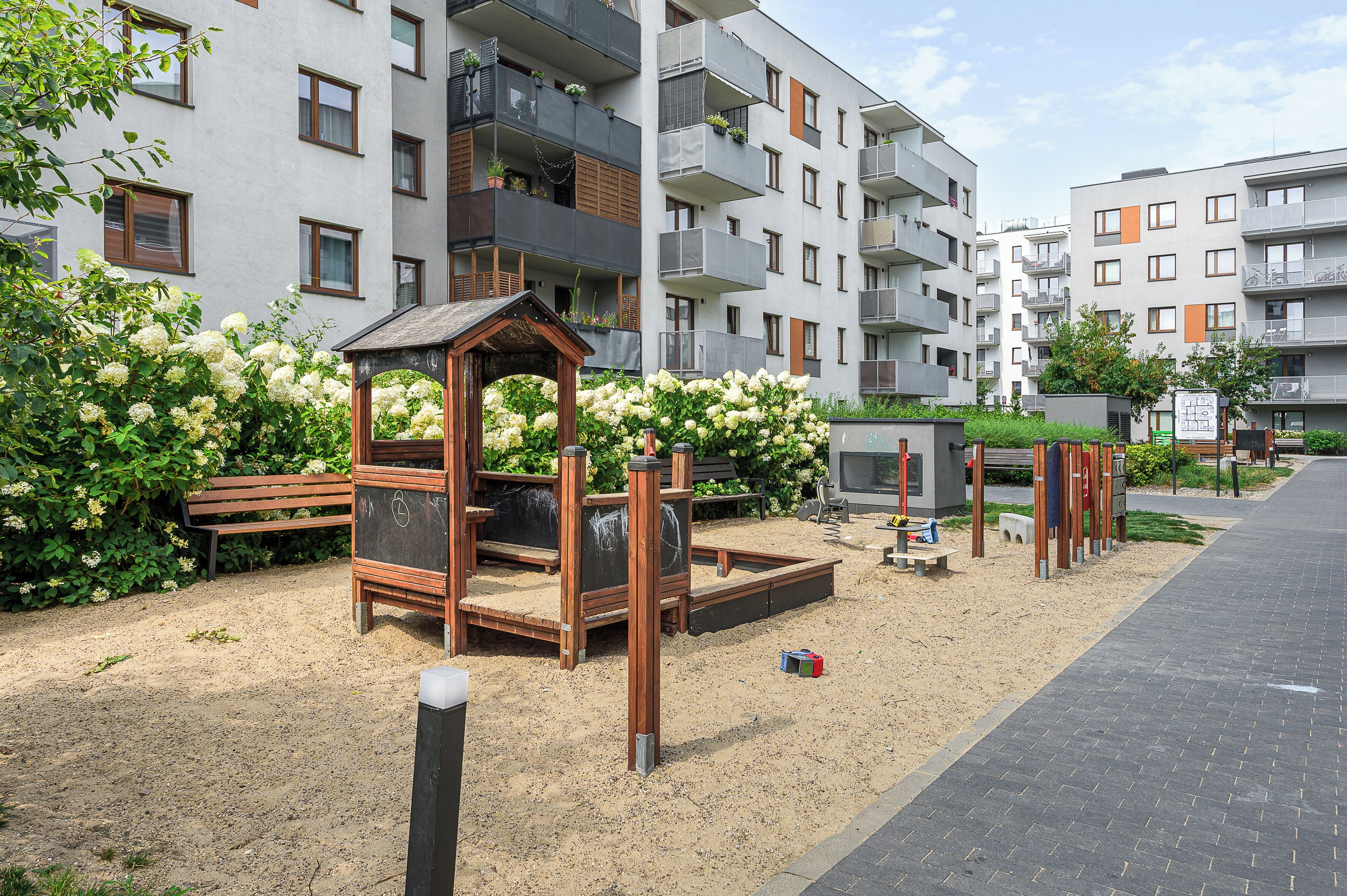 Un terrain de jeux avec une maisonnette en bois et un bac à sable, entouré de verdure et d’immeubles d’habitation.