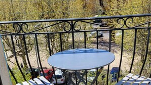 Ein Balkon mit Tisch und Stühlen, eine Fußmatte mit dem Aufdruck 'Welcome Home' und Blick auf Bäume sowie parkende Autos.