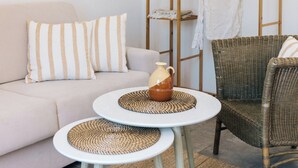 Un espacio habitable minimalista con un sofá, dos mesas redondas, un sillón de mimbre, un estante de madera y una alfombra tejida.