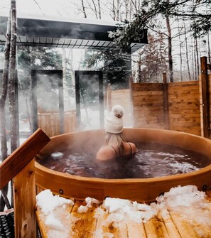 A hot tub outdoors with a person in it, surrounded by snow and wooden structures.