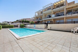 A swimming pool with a tiled deck, a wooden fence, and a building with balconies in the background. - Deluxe Apartment with Pool, Sea View, Shared Pool, and Air Conditioning (Paduledda)