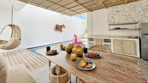 A dining area with a wooden table set for breakfast, a hanging chair, and a stone wall with a bear decoration. - Villa Lavana Uluwatu (Badung)