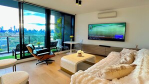 A modern living room with a large TV, comfortable seating, and a balcony with a city view. - CASA DELUXE #5 - Pool Gym Sauna Spa City Views Parking (South Yarra)