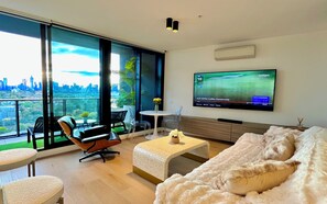 Ein modernes Wohnzimmer mit großem Fernseher, bequemen Sitzgelegenheiten und einem Balkon mit Blick auf die Stadt.