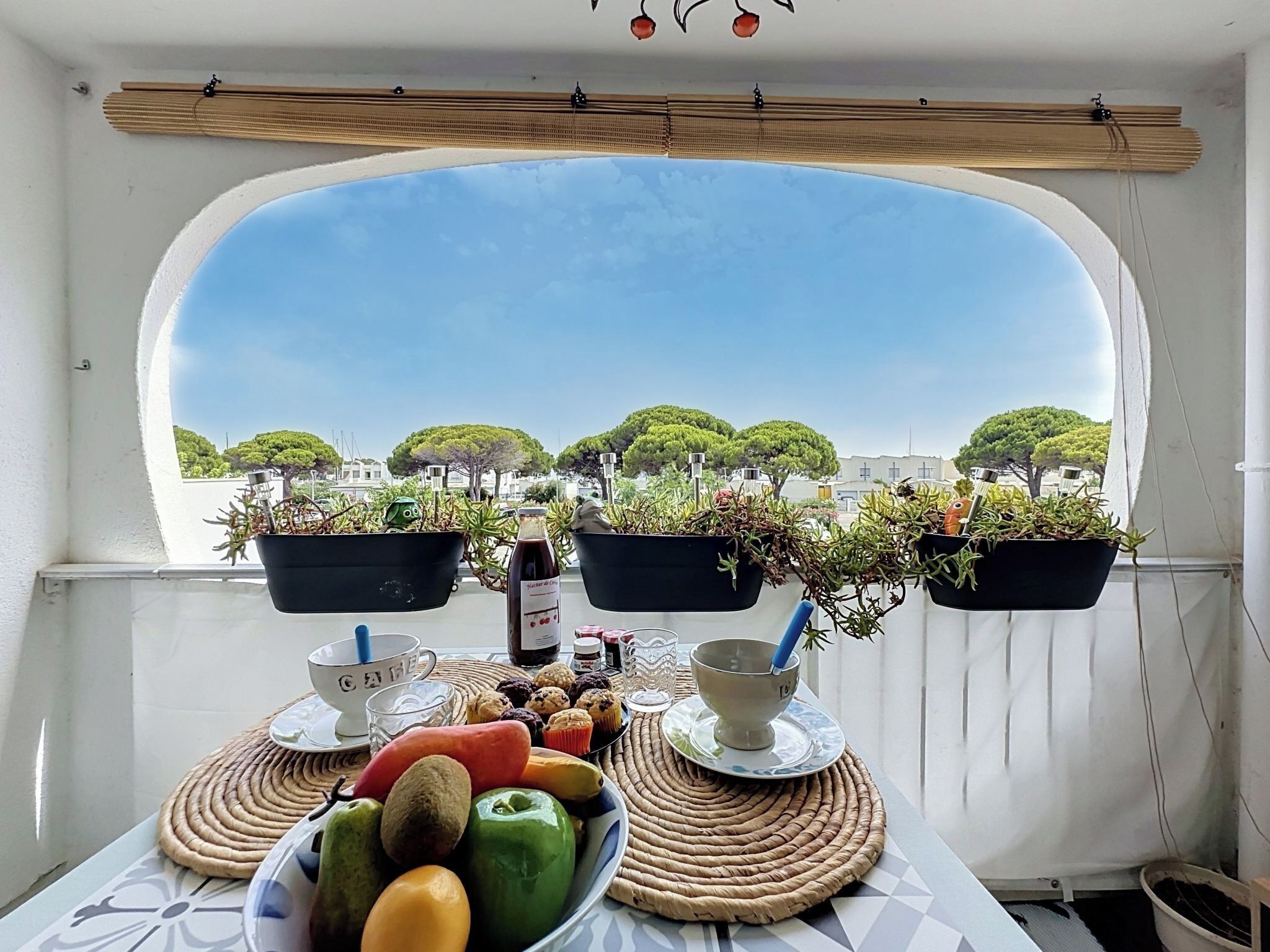 Un balcón con una mesa preparada para una comida, plantas en macetas y vistas a árboles y edificios.