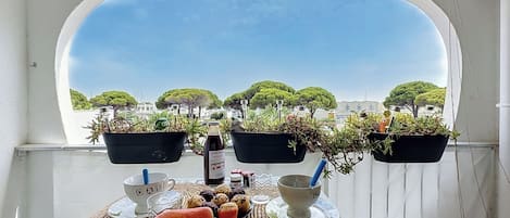 Un balcón con una mesa preparada para una comida, plantas en macetas y vistas a árboles y edificios.