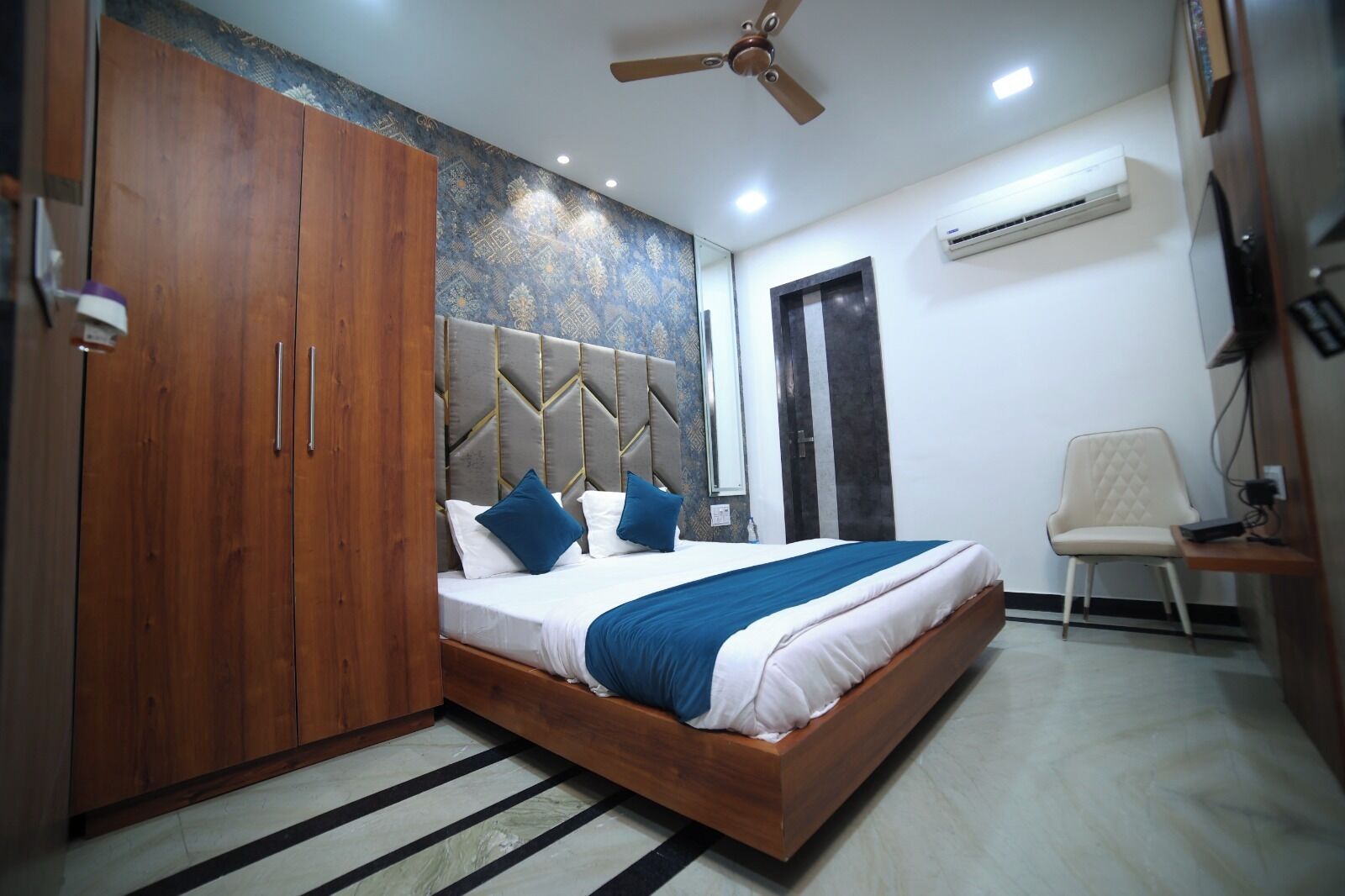 A bedroom with a bed, wooden wardrobe, ceiling fan, air conditioning unit, and a chair.