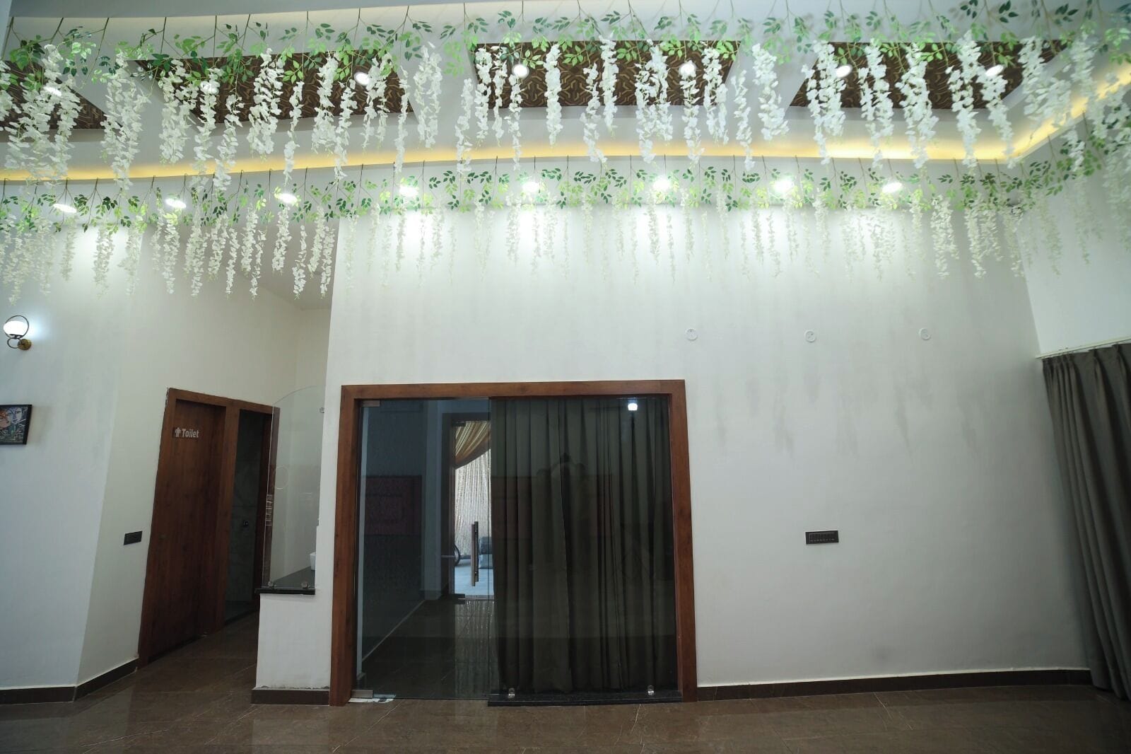 A hallway with a wooden door labeled 'Toilet,' a glass door, and hanging artificial white flowers.