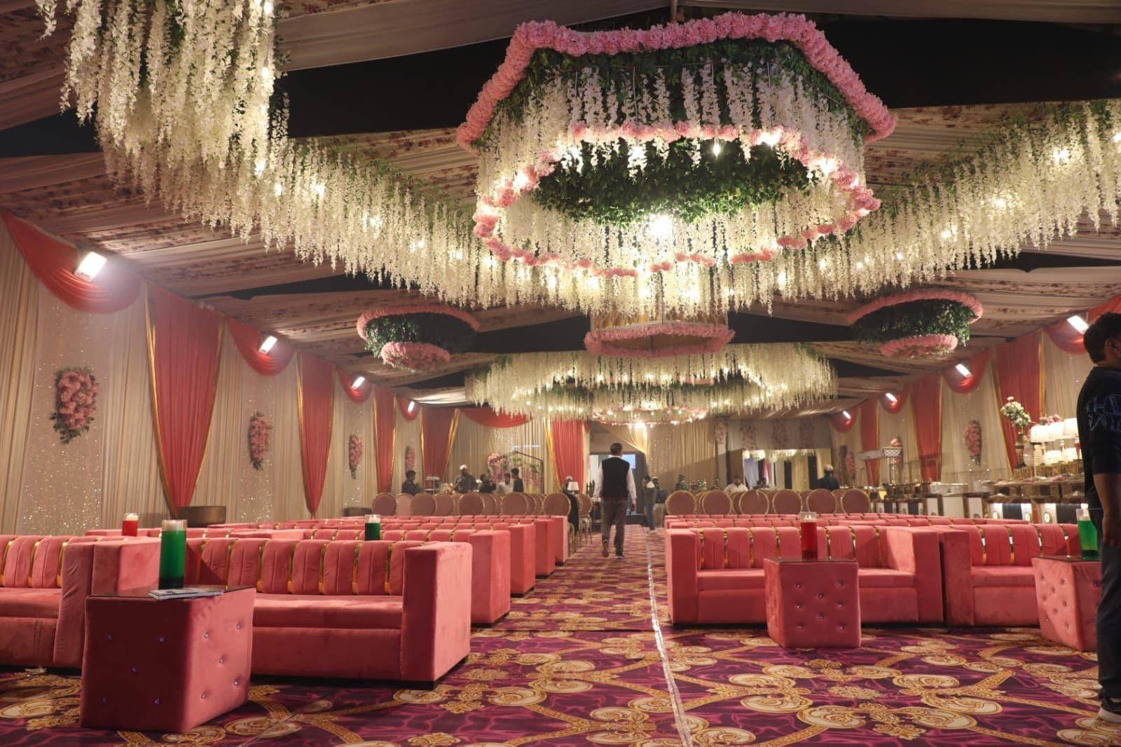 A banquet hall with rows of red upholstered sofas, a patterned carpet, and hanging floral decorations.