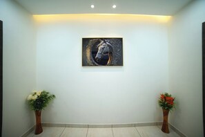 A hallway with a framed artwork on the wall, two potted plants, and a light-colored floor. - HOTEL ROYAL PALACE (Bhiwani)