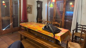 Un coin repas avec une table en bois, des bancs et des chaises. Il y a une horloge au mur et une fenêtre avec des rideaux.