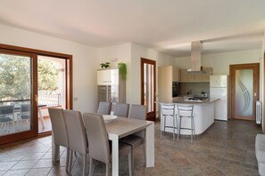 A modern kitchen with a central island, dining area with chairs, and a large sliding glass door leading to an outdoor patio. - Villa Certosina by Wonderful Italy (Siniscola)