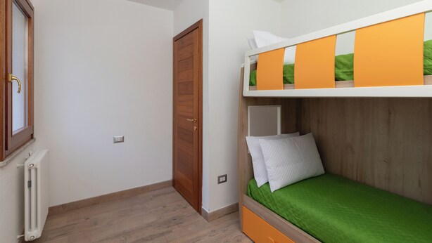 Una stanza con un letto a castello, una porta in legno, un termosifone e una finestra.