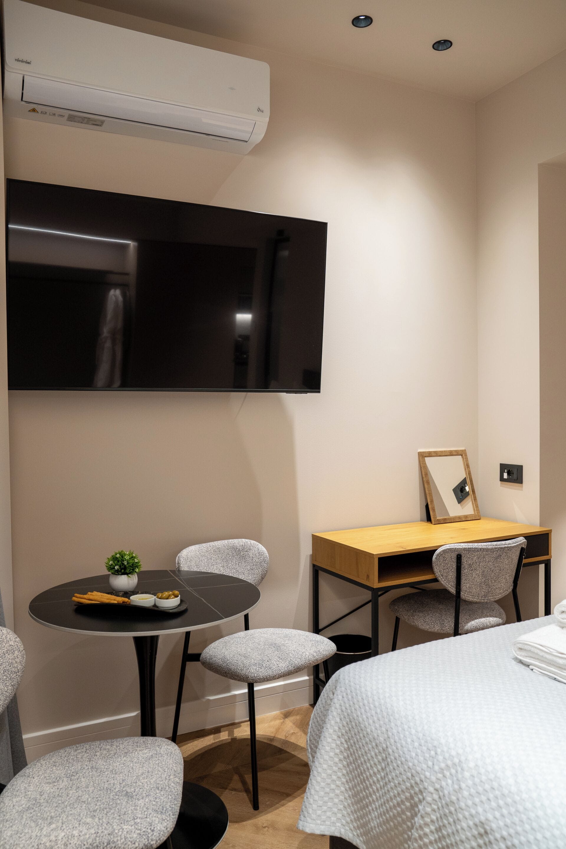 A modern hotel room with a flat-screen TV mounted on the wall, a small round table with a plant and a bowl of olives, two grey upholstered chairs, a wooden desk with a mirror, and a bed with a white duvet.