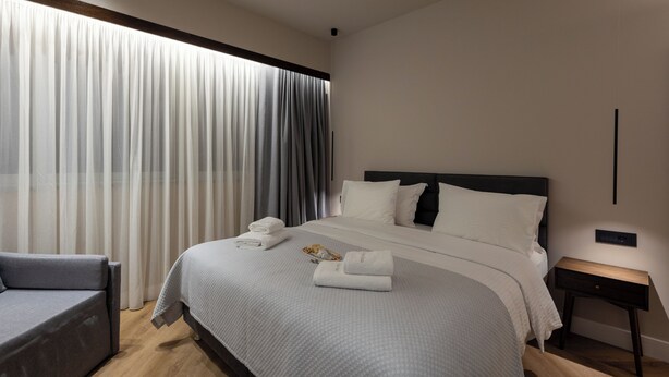 A modern hotel room with a large bed, a bedside table, a chair, and a window with curtains. - Lux Suites Athens (Athina)