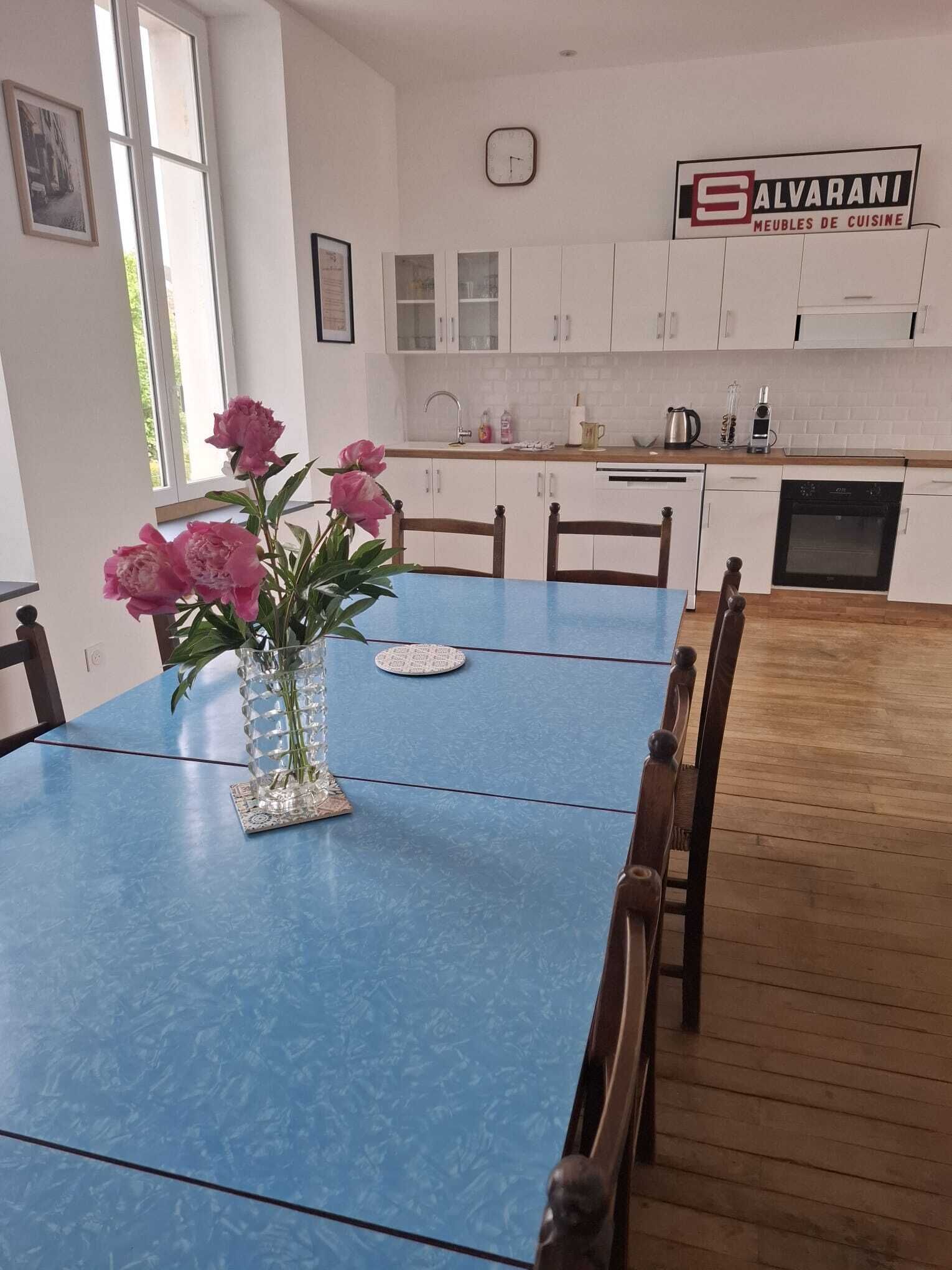 Une table à manger avec des chaises, un vase de fleurs, une horloge et un coin cuisine avec des placards et des appareils électroménagers.