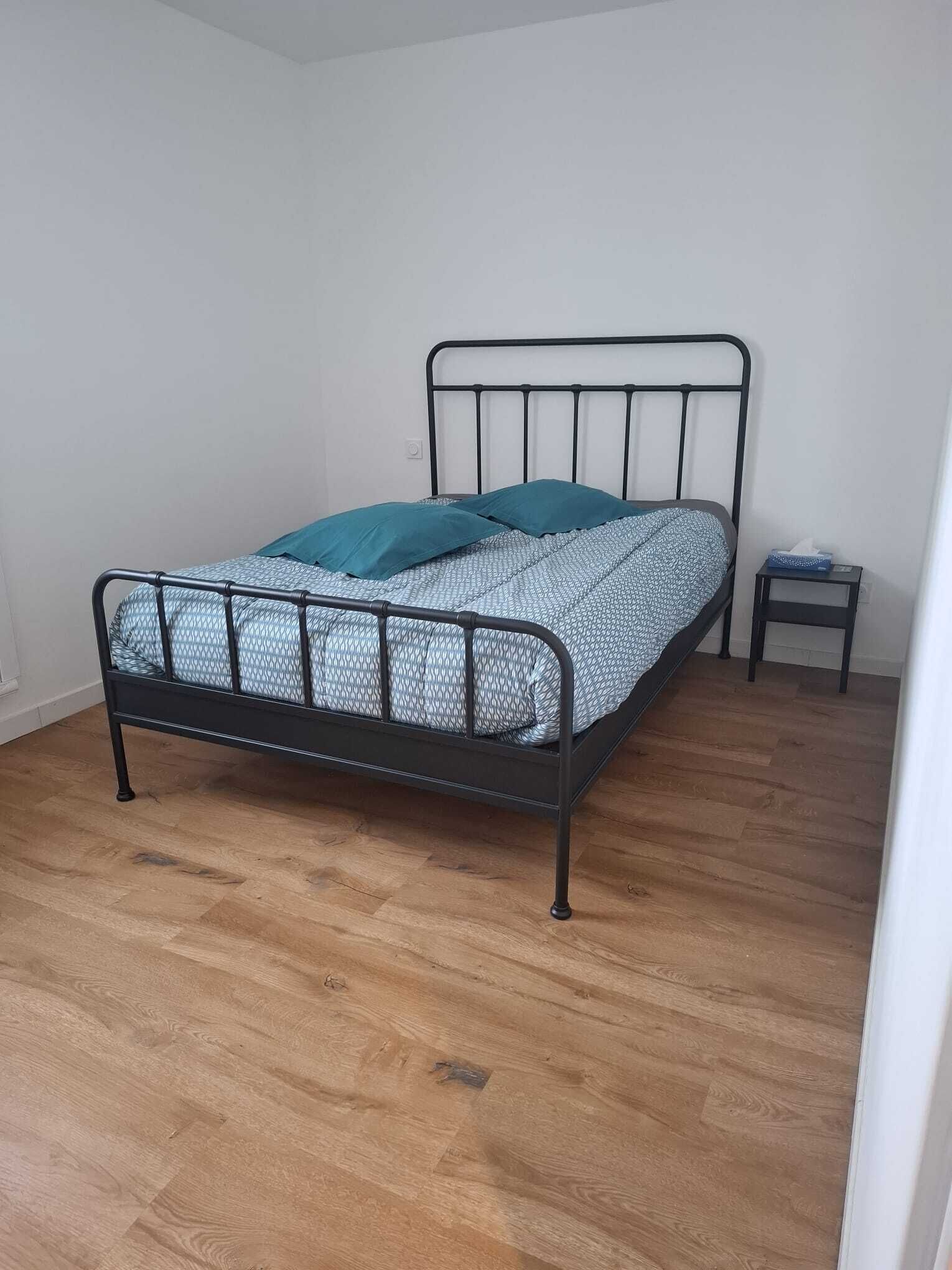 En säng med metallram, ett mönstrat överkast och en turkosfärgad kudde. Bredvid sängen står ett litet sidobord med en näsduksask. Rummet har trägolv och vita väggar.