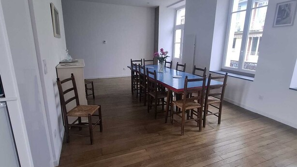 A dining area with a wooden table and chairs, a small kitchen area with a refrigerator, and large windows providing natural light.