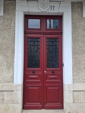 A red double door with intricate metalwork, a white door frame, and a stone wall with a number plaque above. - 200-square-meter apartment - 5 bedrooms - 3 bathrooms (Avallon)