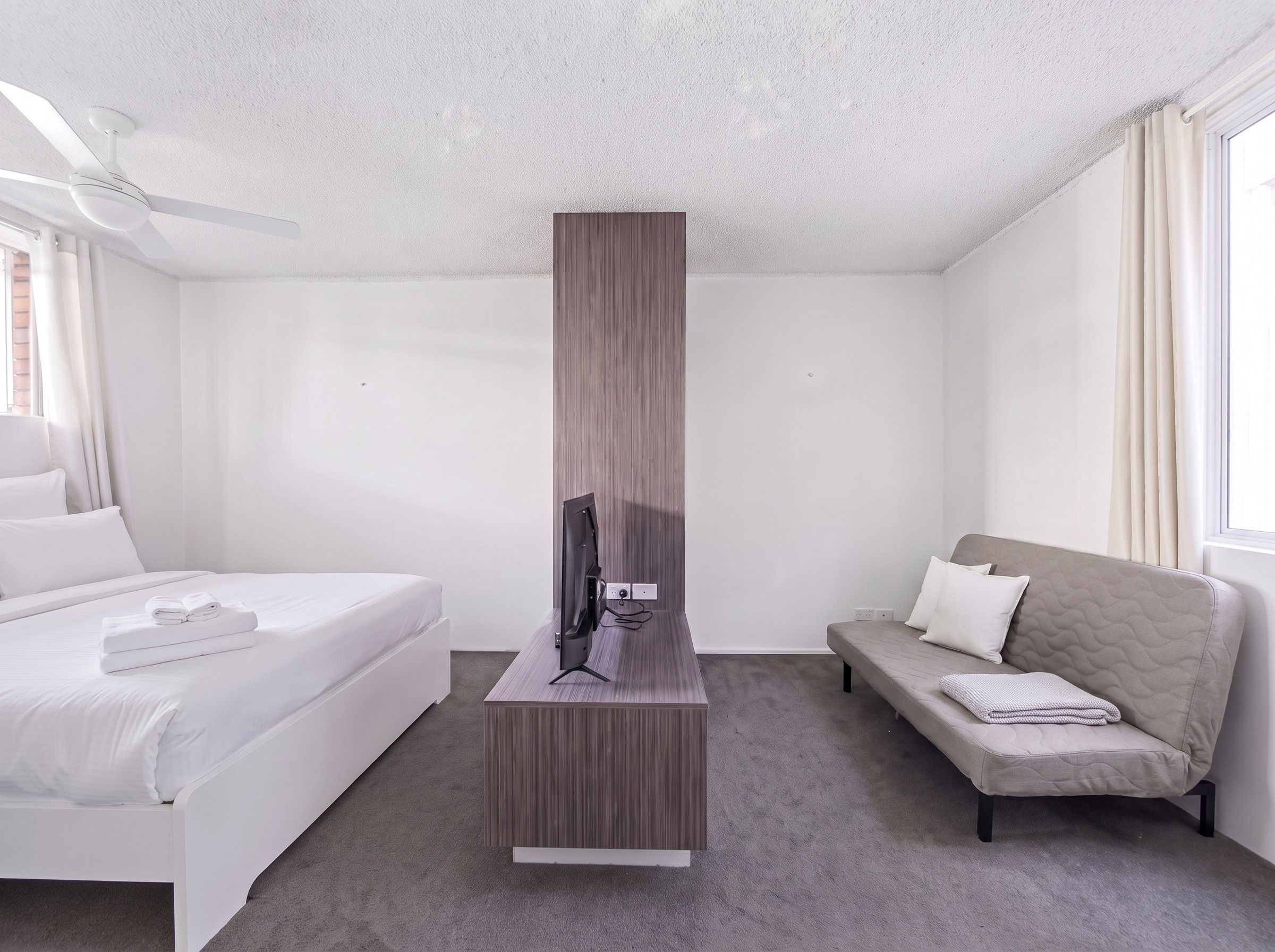A hotel room with a bed, a sofa, a flat-screen TV on a stand, and a ceiling fan.