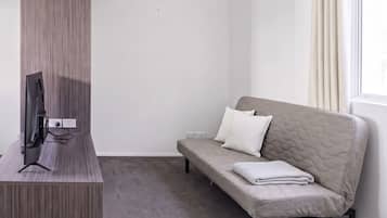 A room with a flat-screen TV on a wooden stand, a grey sofa with cushions, and a window with curtains.